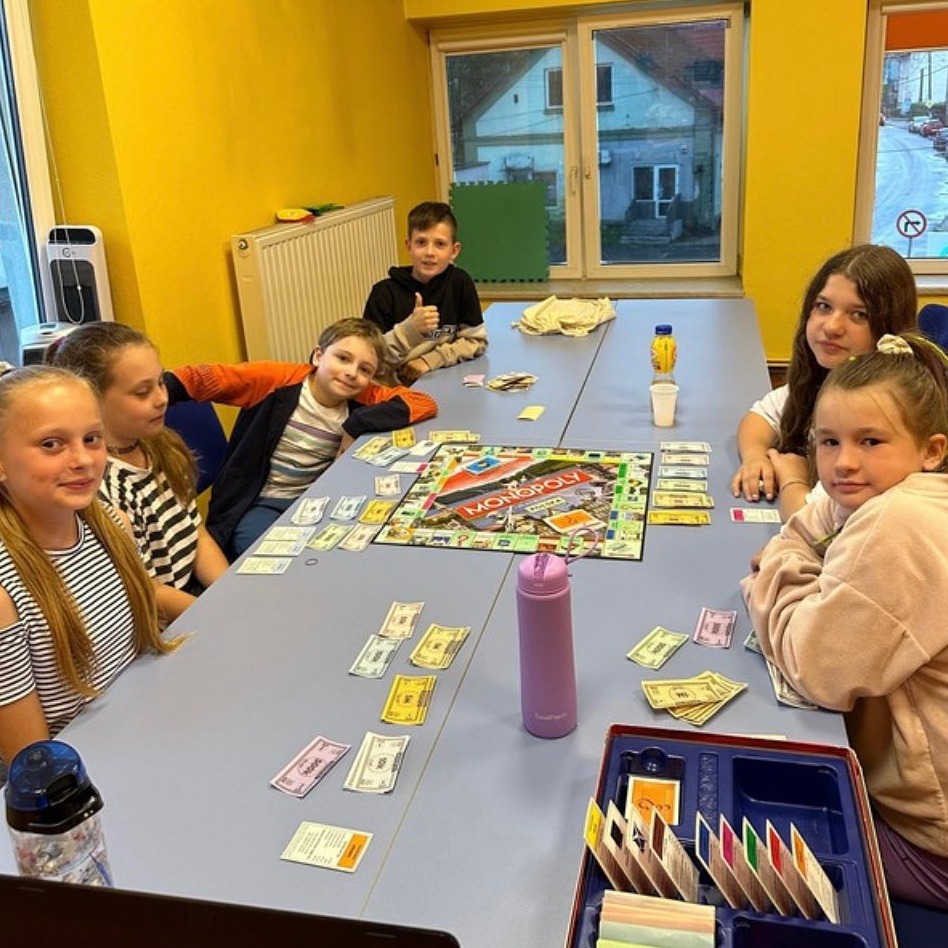 Grupa dzieci w jasnym pomieszczeniu siedzi wokół stołu grając w Monopoly. Na stole rozłożona plansza gry, karty i banknoty. Jeden z chłopców pokazuje kciuk w górę.