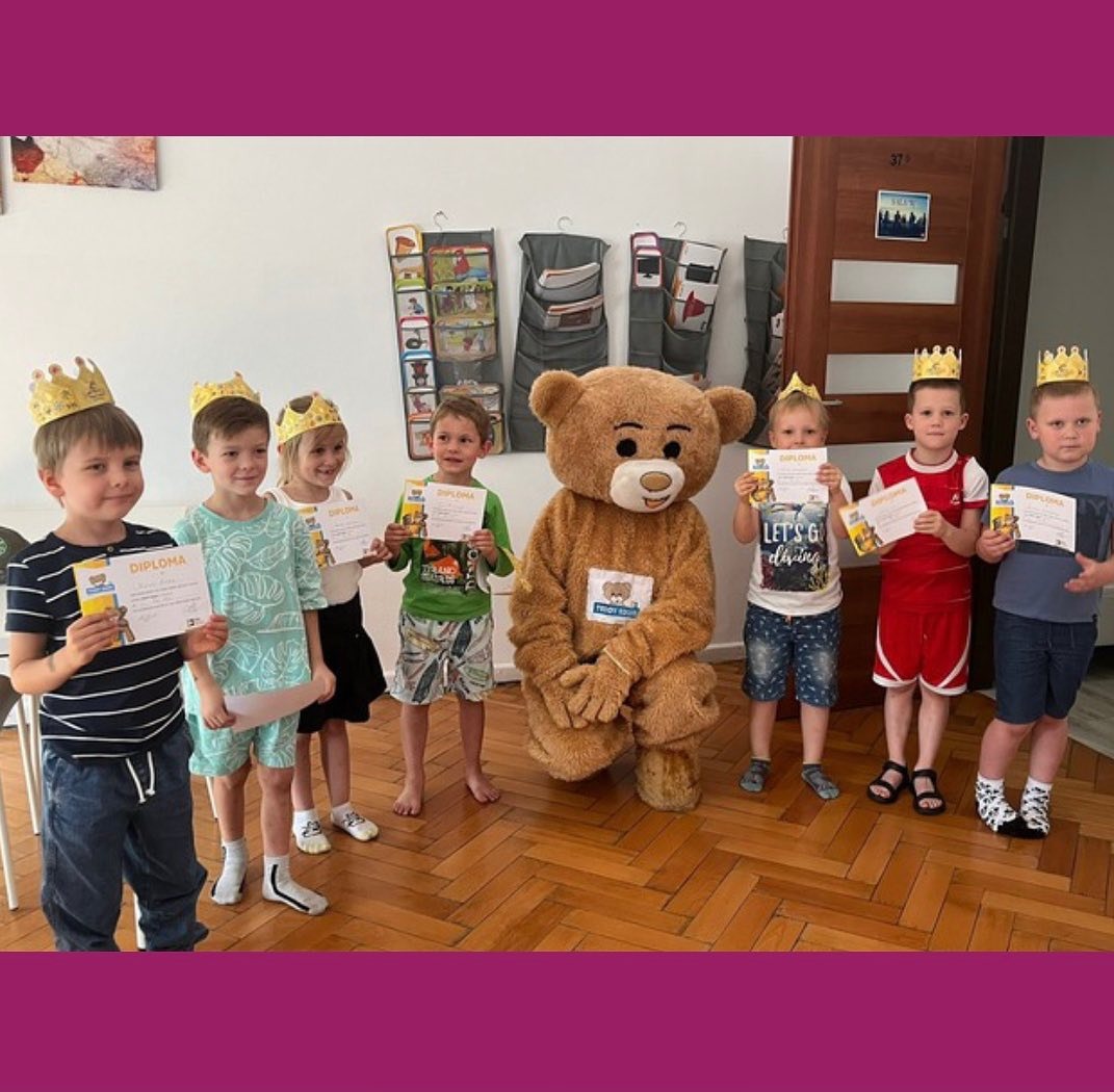Grupa dzieci w koronach trzyma dyplomy, obok nich stoi osoba przebrana za pluszowego misia z logo firmy na kostiumie, w tle ściana z organizerami i drzwi z naklejką.