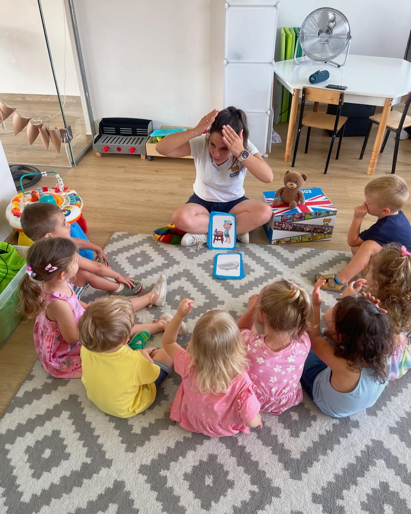 Nauczycielka siedzi na podłodze z grupą dzieci, używając kart obrazkowych do nauki słówek, w tle zabawki i meble w jasnym pomieszczeniu.