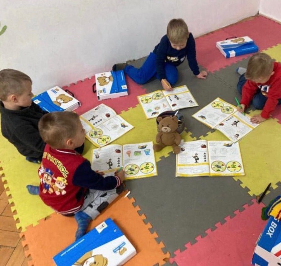 Czterech małych chłopców siedzi na podłodze z puzzli, ucząc się z otwartych książek z rysunkami, obok nich leżą pudełka z misiem i pluszowy miś