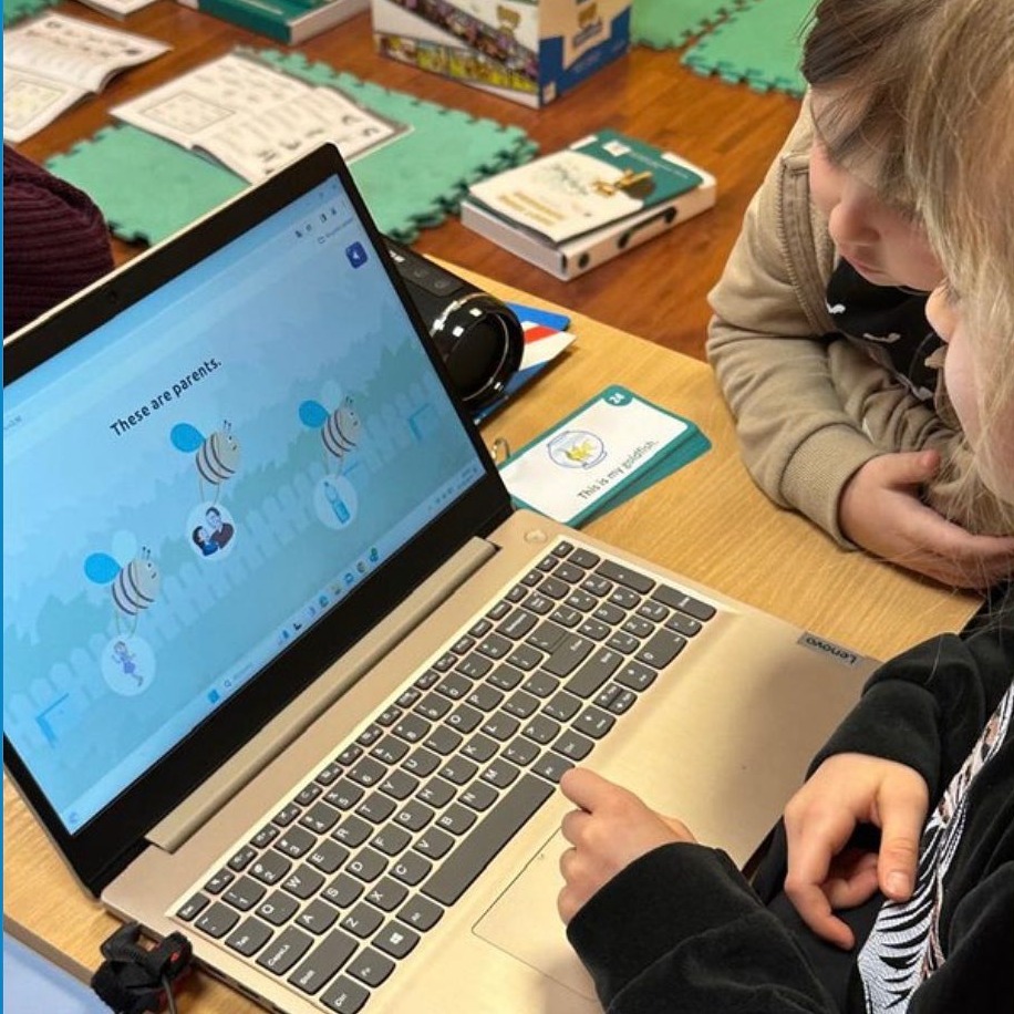 Dzieci patrzą na ekran laptopa z interaktywną lekcją angielskiego, na biurku leżą karty edukacyjne i inne pomoce naukowe.