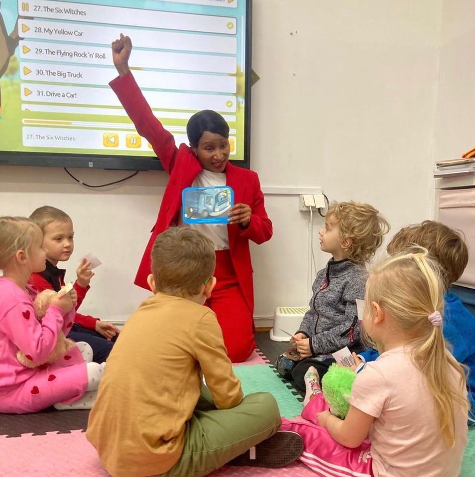 Nauczycielka w czerwonym garniturze, klęcząca przed grupą dzieci siedzących na podłodze, pokazuje ilustrację niebieskiej ciężarówki, z interaktywną tablicą wyświetlającą listę piosenek w tle.