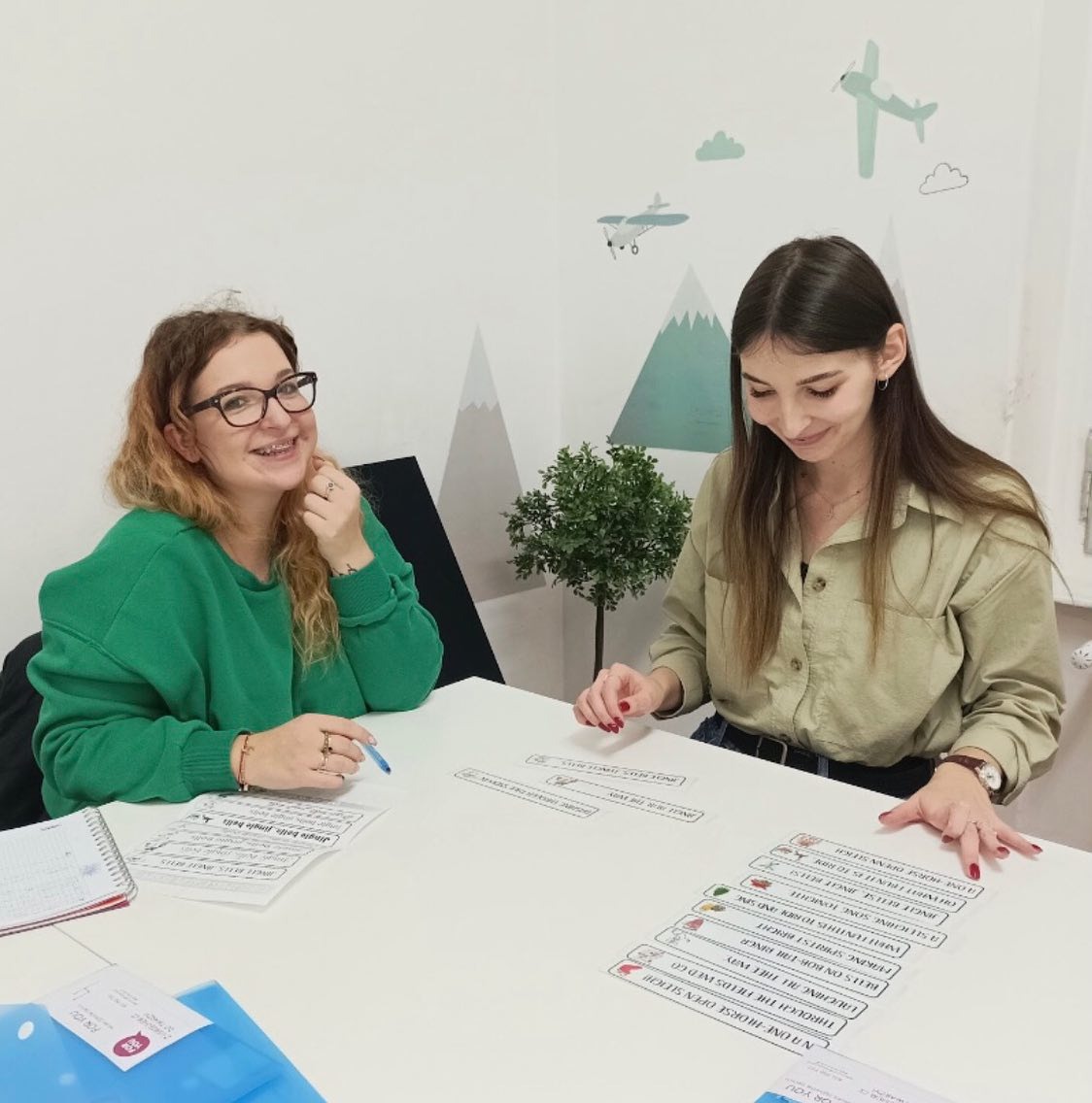 Dwie młode kobiety siedzą przy stole podczas lekcji języka angielskiego, jedna z nich uśmiecha się do kamery, na stole rozłożone są karty z angielskimi zwrotami, w tle ściana z dekoracjami...