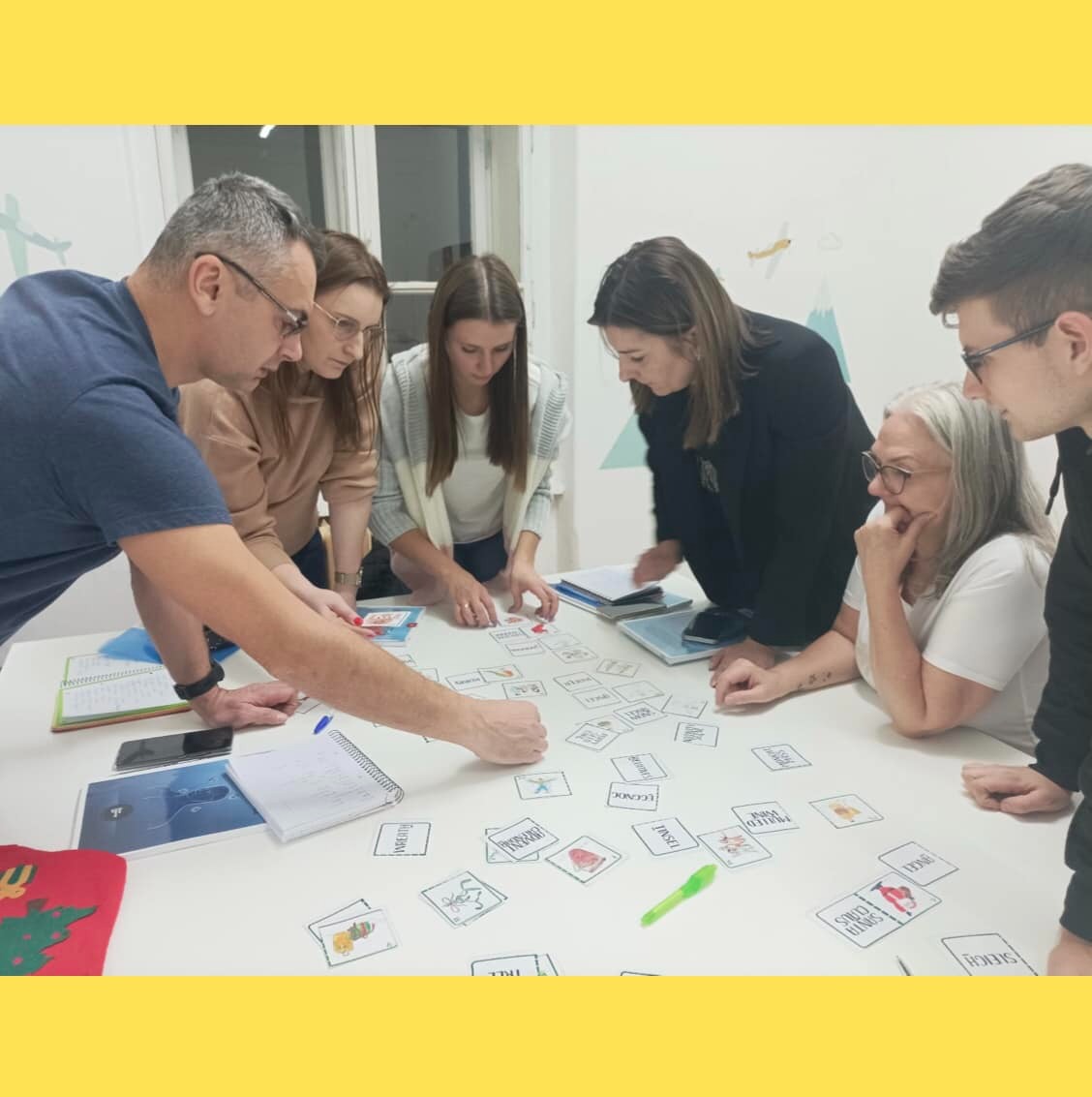 Grupa osób skupiona wokół stołu z kartami obrazkowymi podczas lekcji językowej, notatniki i długopisy na blacie.
