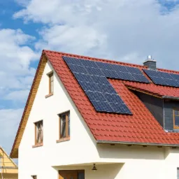 Dom jednorodzinny z czerwoną dachówką i panelami słonecznymi na dachu, widoczny w tle budynek w trakcie budowy z rusztowaniami.