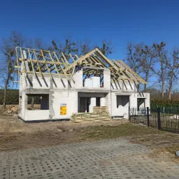 Wznoszenie więźby dachowej na surowym budynku z betonu komórkowego w słoneczny dzień, widoczne drewniane elementy konstrukcyjne i paczka desek na placu budowy.