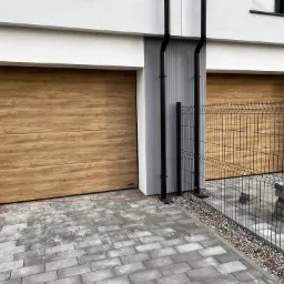 Dwie segmentowe, drewnopodobne bramy garażowe z panelami poziomymi, zamontowane w nowoczesnym budynku z szarą elewacją, widoczne z zewnątrz przez czarne metalowe ogrodzenie.