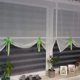 Dwa okna w Gdańsku z roletami w biało-szare pasy, upięte zwiewną, białą firanką z zielonymi kokardkami, na parapecie rośliny doniczkowe i zdjęcia w ramkach, obok kryształowy żyrandol.