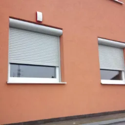 Dwa okna z opuszczonymi, szarymi roletami zewnętrznymi na pomarańczowej ścianie budynku, widoczny fragment balkonu z szarą balustradą.