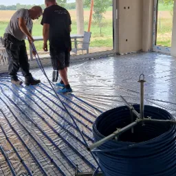 Montaż ogrzewania podłogowego: dwóch instalatorów rozkłada niebieskie rury na folii z nadrukiem siatki, widoczna rolka rur na metalowym stojaku, duże okna w tle.