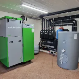 Kotłownia z zainstalowanym piecem na paliwo stałe SAS z zielonymi akcentami, zbiornikiem akumulacyjnym, grupą pompową i rozbudowaną instalacją rur w piwnicy z kafelkami na podłodze.