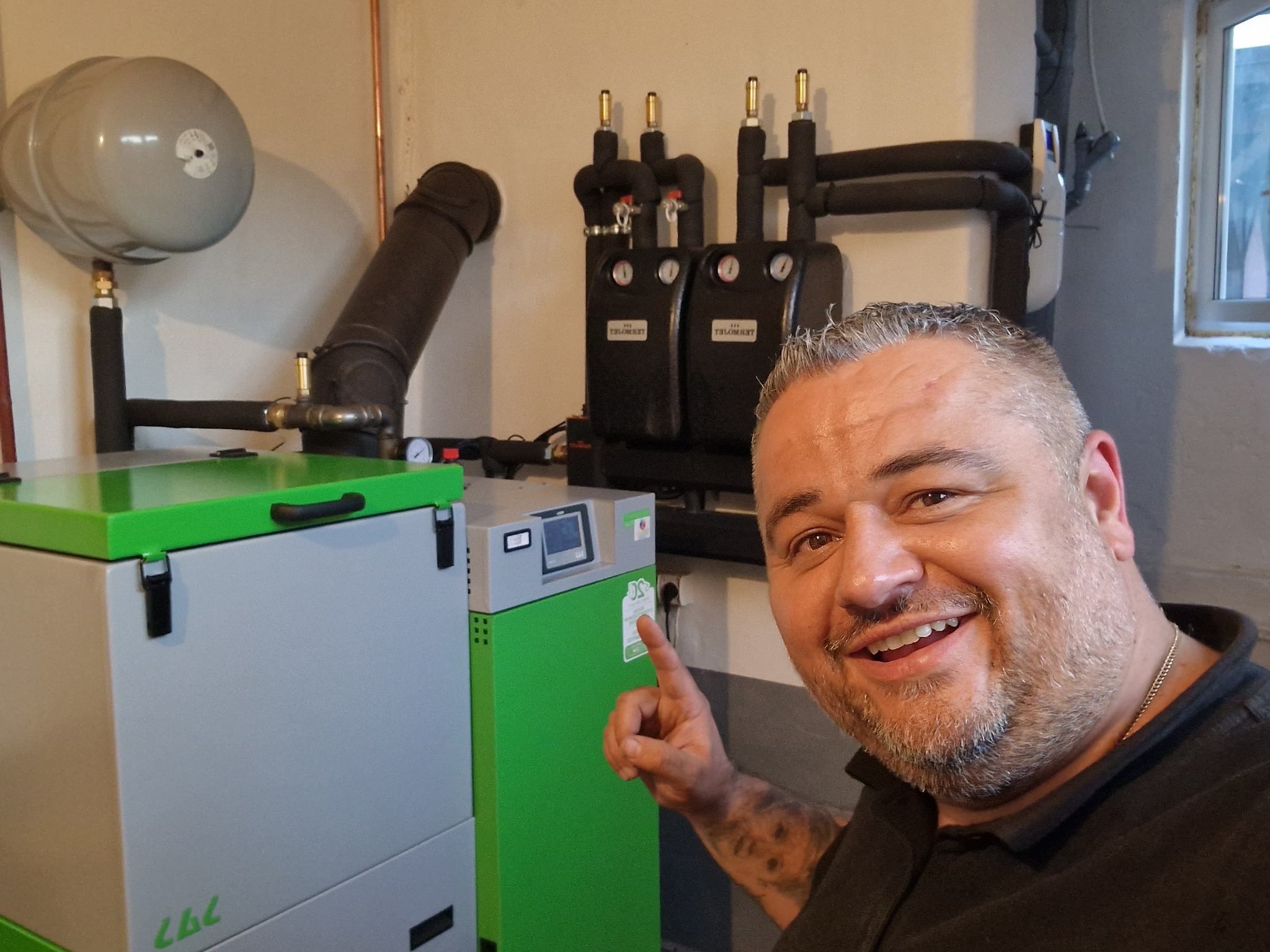 Uśmiechnięty mężczyzna wskazuje na nowoczesny piec na paliwo stałe z zielonymi akcentami, zainstalowany w kotłowni z widoczną instalacją grzewczą.