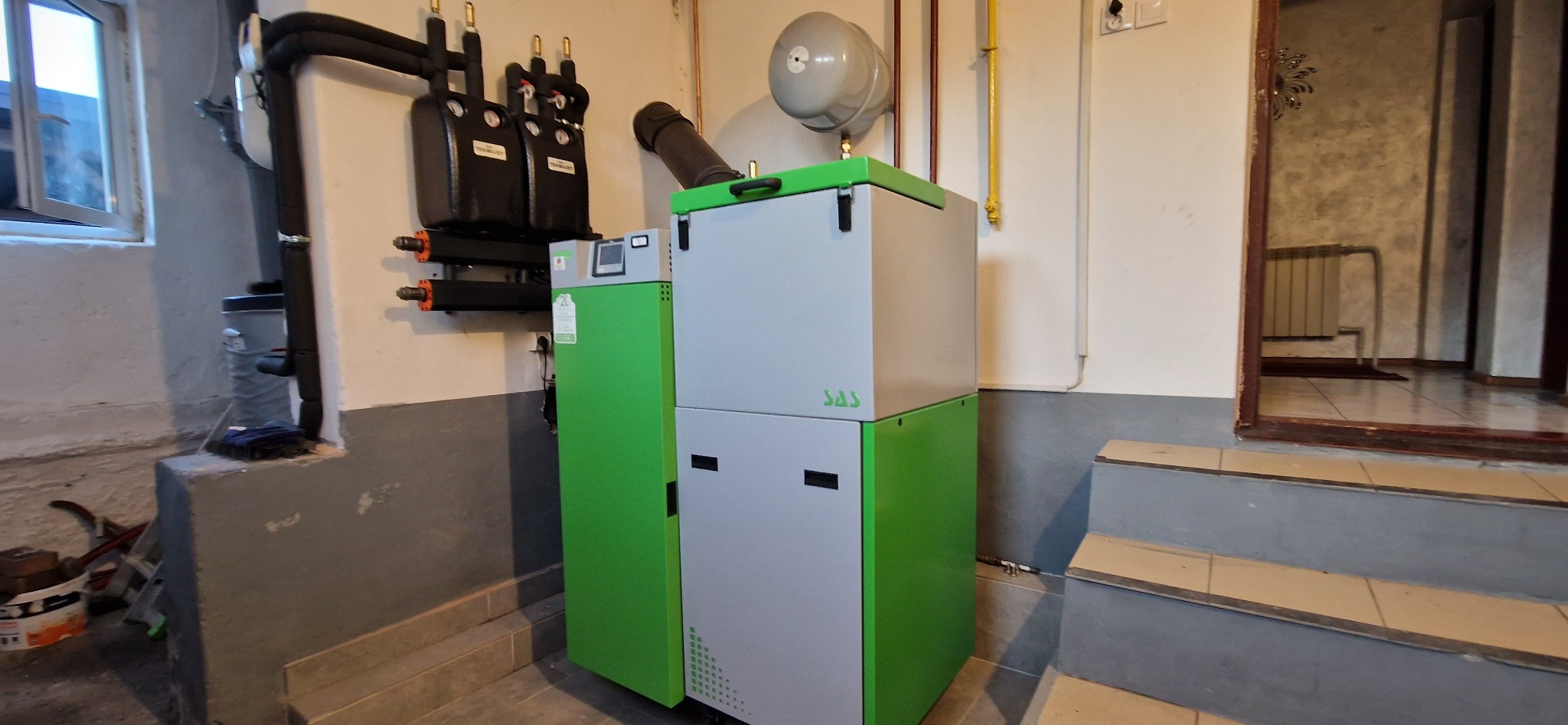 Nowoczesny, dwukomorowy piec na paliwo stałe SAS w kotłowni, z widocznymi elementami instalacji i schodami prowadzącymi do pomieszczenia.