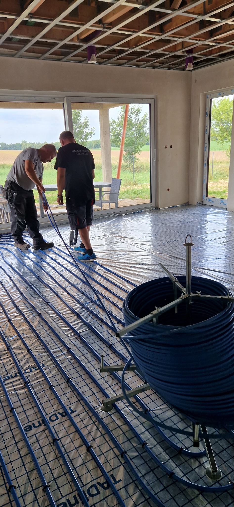 Montaż ogrzewania podłogowego: dwóch instalatorów rozkłada niebieskie rury na folii z nadrukiem siatki, widoczna rolka rur na metalowym stojaku, duże okna w tle.