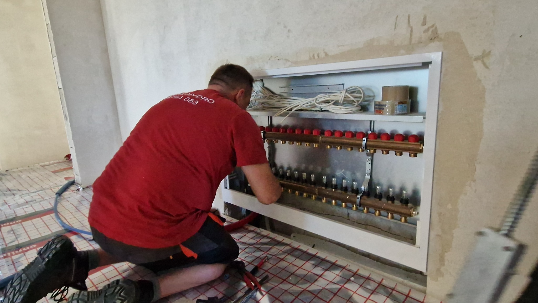 Instalator w czerwonej koszulce montuje rozdzielacz do ogrzewania podłogowego w szafce, widoczna siatka grzewcza na podłodze i narzędzia.