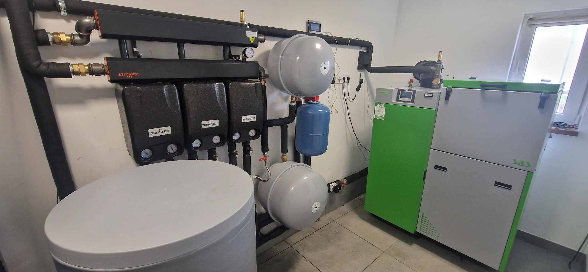 Kotłownia z piecem na paliwo stałe, trzema czarnymi modułami TERMOJET, zbiornikiem buforowym, szarymi zbiornikami wyrównawczymi i orurowaniem w pomieszczeniu z oknem.