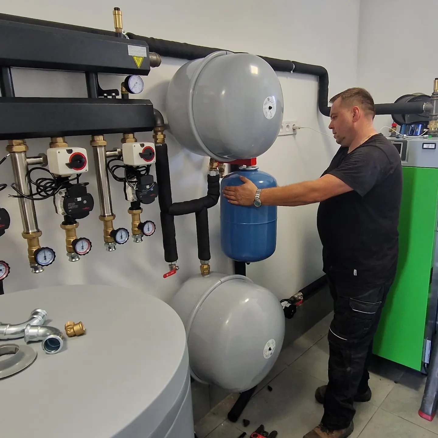 Instalator w czarnym stroju łączy niebieski zbiornik wyrównawczy do instalacji grzewczej z szarymi zbiornikami, widoczne manometry, pompy obiegowe i zielony piec grzewczy.