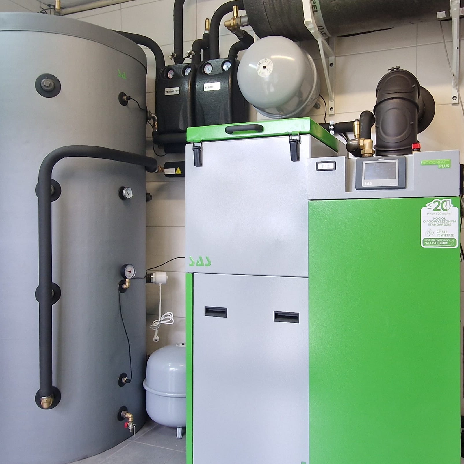 Nowoczesny kocioł na paliwo stałe Bio Compact Plus marki SAS, z szarym zbiornikiem buforowym i czarnymi rurami instalacji grzewczej, zamontowany w kotłowni z białymi płytkami.
