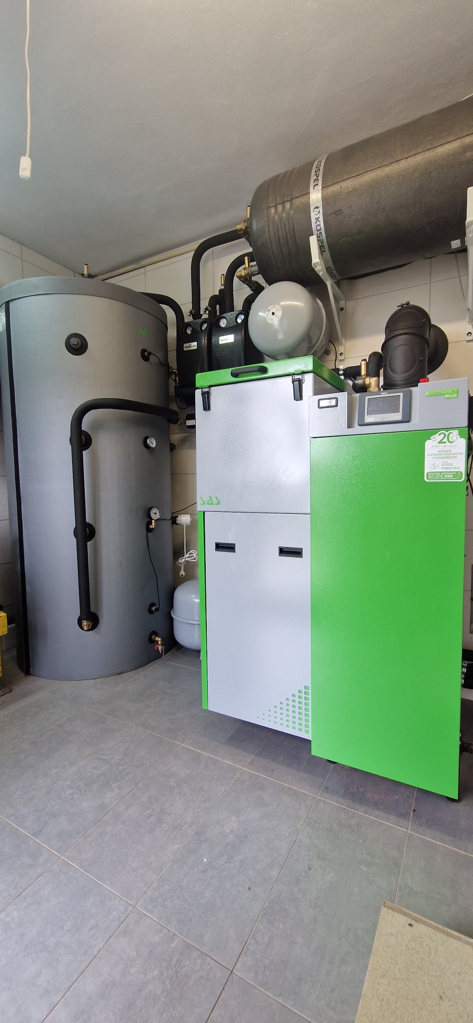 Instalacja kotła na paliwo stałe SAS Bio Compact iplus z zasobnikiem, zbiornikiem buforowym Kospel i osprzętem hydraulicznym w kotłowni wyłożonej szarą terakotą.