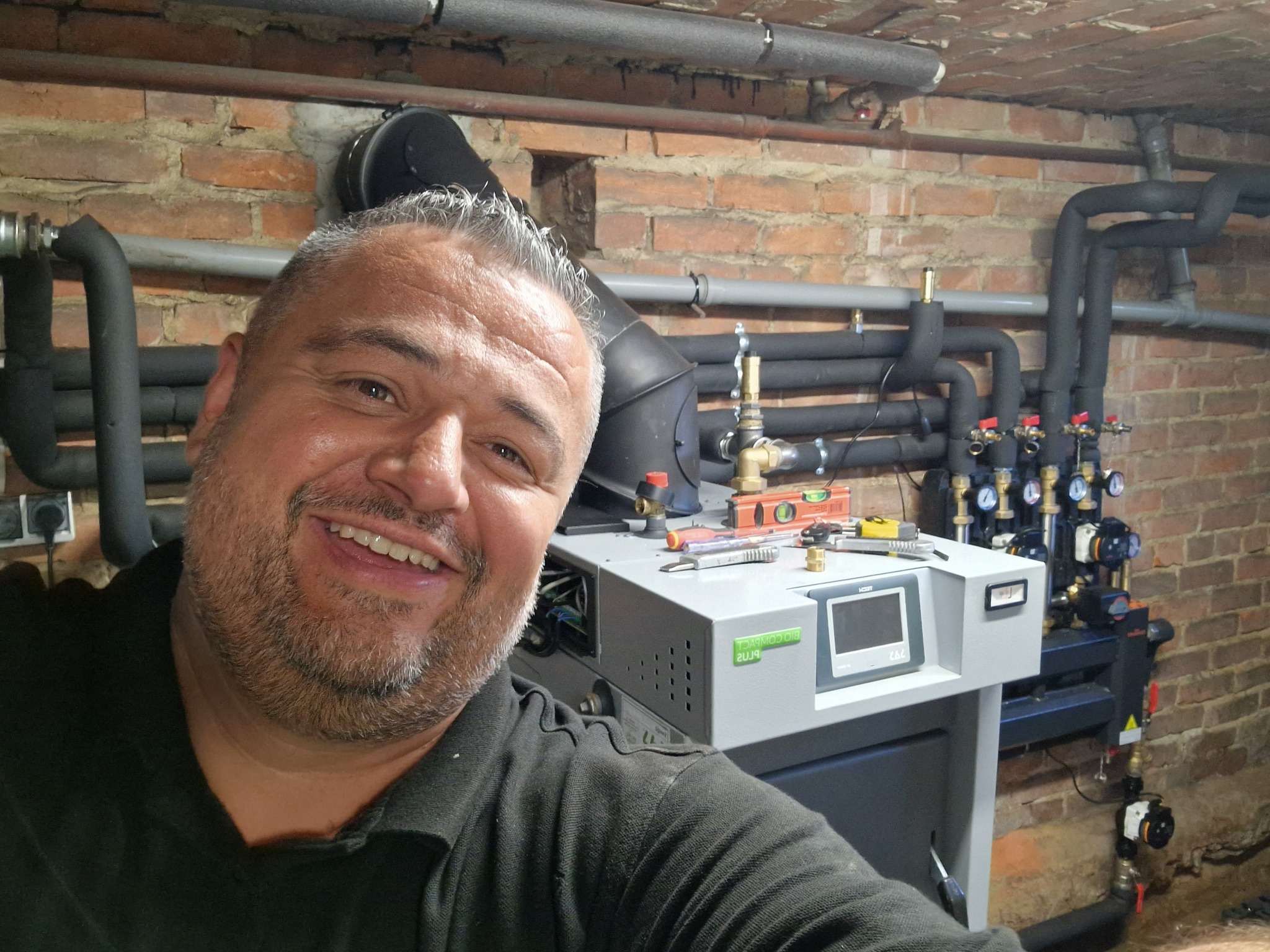 Uśmiechnięty mężczyzna pozuje przed nowym kotłem grzewczym Bio Compact Plus, z widocznymi narzędziami, rurami i zaworami na tle ceglanej ściany w kotłowni.