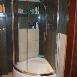 Kabina prysznicowa narożna z chromowanymi profilami, półkami z kosmetykami i ciemnymi, wzorzystymi płytkami na ścianach, uzupełniona o jasne płytki i akcesoria łazienkowe.