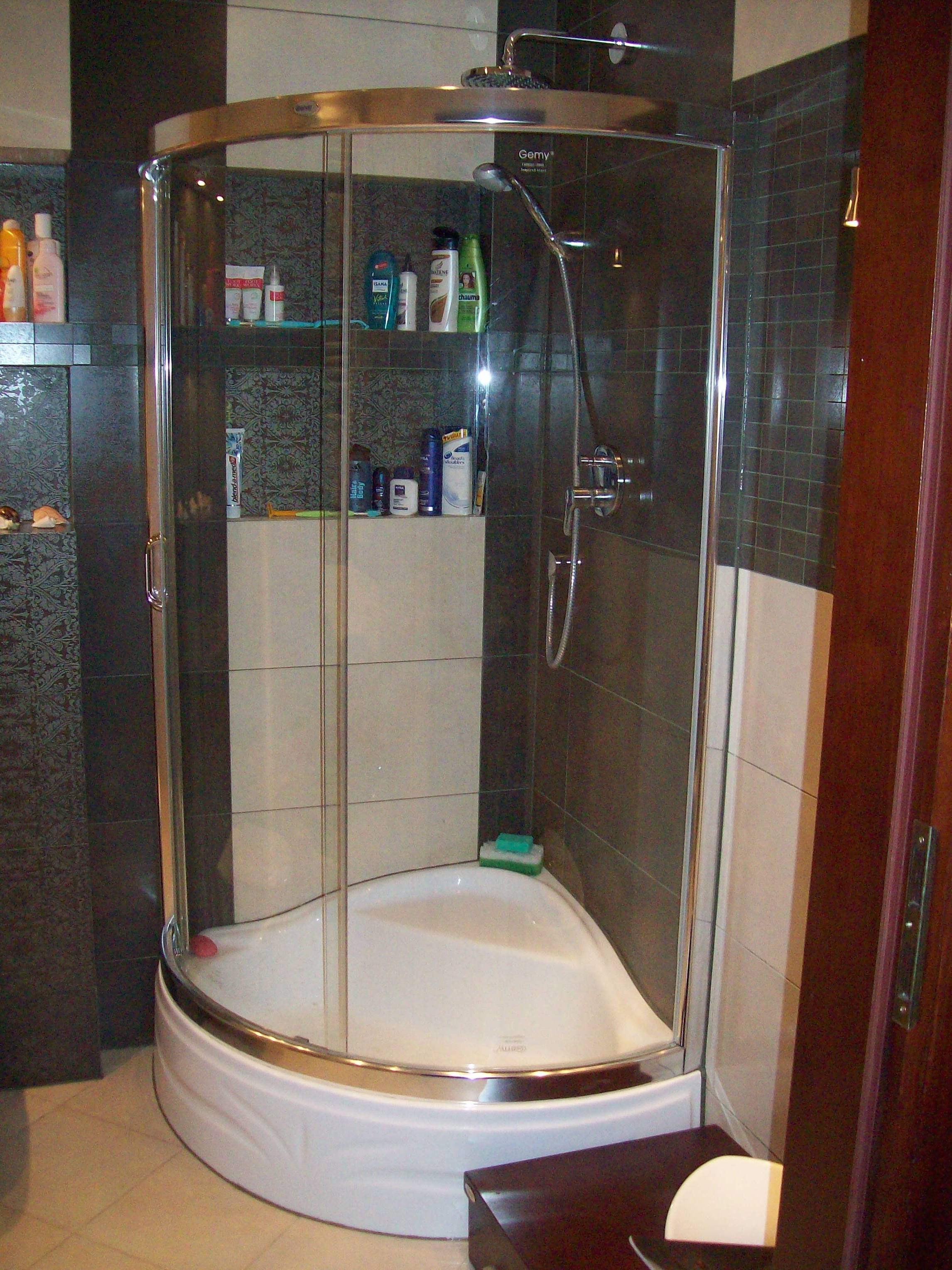 Kabina prysznicowa narożna z chromowanymi profilami, półkami z kosmetykami i ciemnymi, wzorzystymi płytkami na ścianach, uzupełniona o jasne płytki i akcesoria łazienkowe.