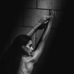 Czarno-białe zdjęcie kobiety w ozdobnej sukni, opierającej się rękami o kamienną ścianę w dramatycznym oświetleniu.