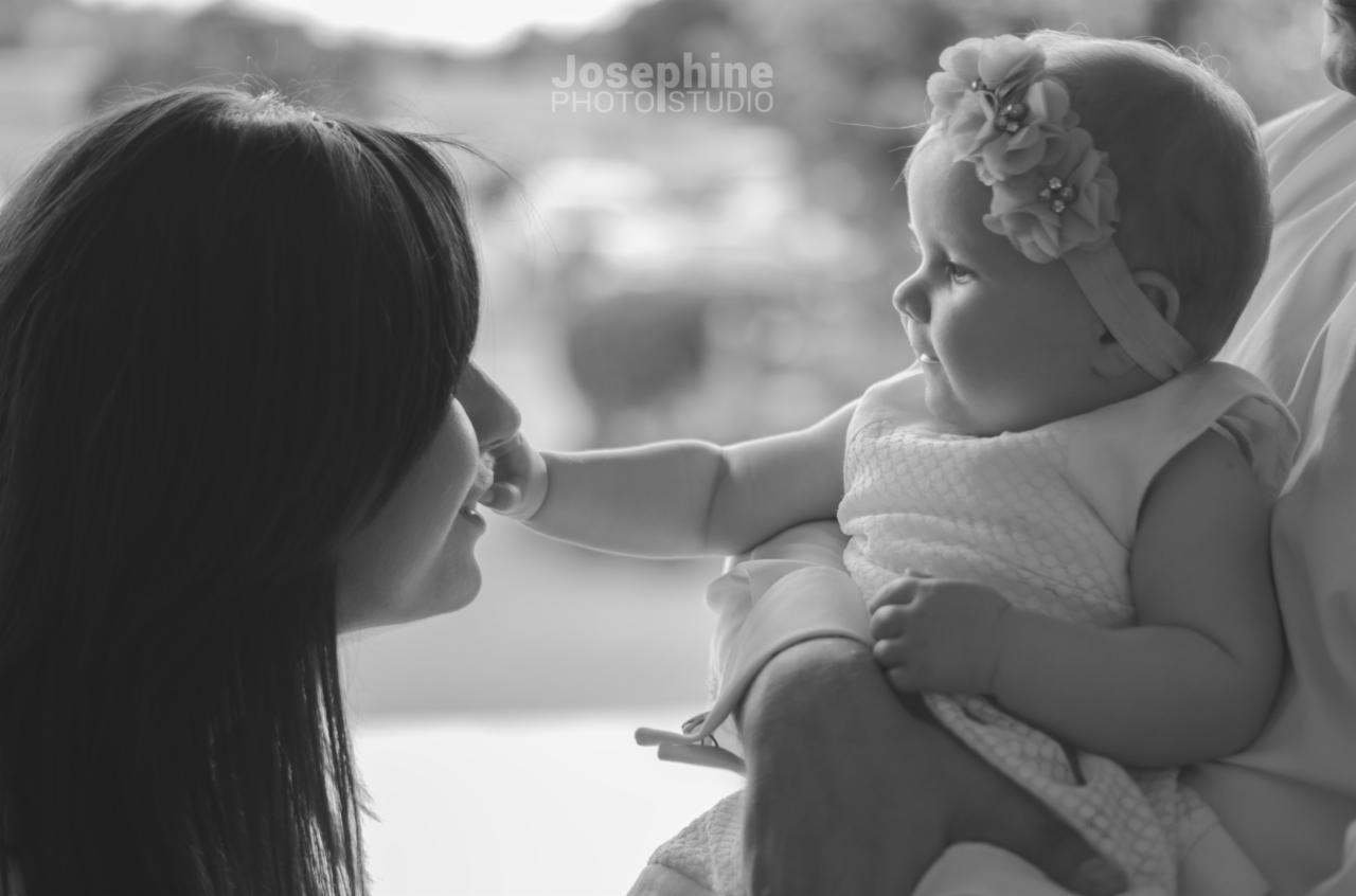 Czarno-białe zdjęcie przedstawia mamę, której dziecko w sukience i opasce z kwiatami dotyka nosa.