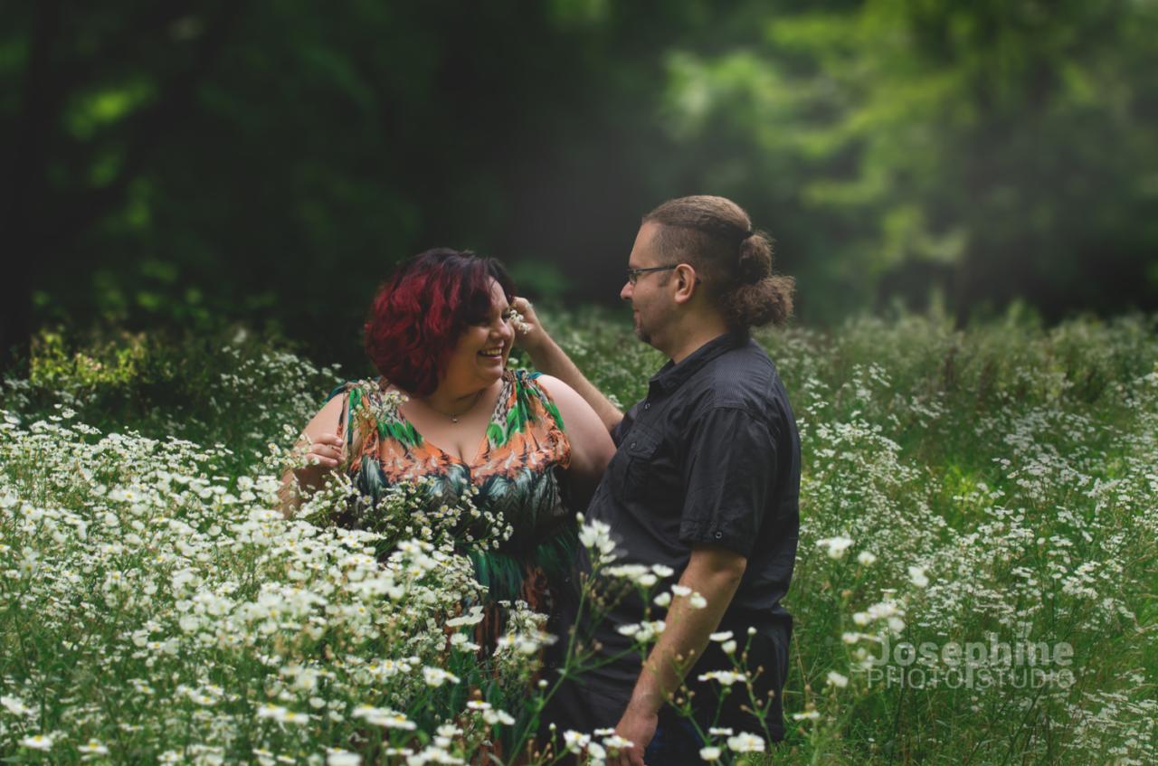Uśmiechnięta para w kwitnącej łące, kobieta z czerwonymi włosami trzyma białe kwiaty, mężczyzna w czarnej koszuli patrzy na nią.