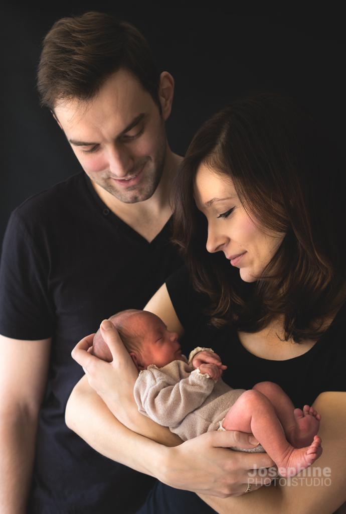 Rodzice spoglądają na śpiącego noworodka w beżowym ubranku, trzymanego w ramionach matki na ciemnym tle.