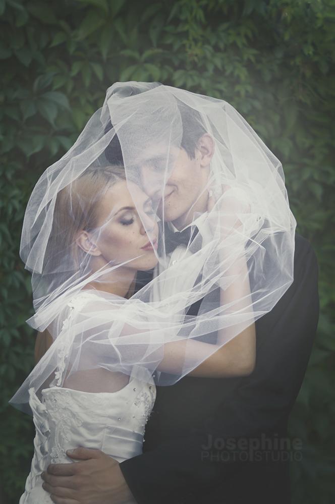 Para młoda w objęciach, zakryta delikatnym, zwiewnym welonem na tle zielonych liści.