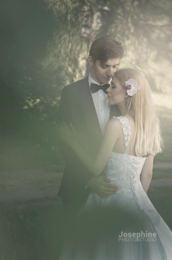 Para młoda w objęciach na tle zamazanego, leśnego tła; panna młoda z blond włosami i orchideą we włosach, w sukni z koronkowym gorsetem, pan młody w czarnym smokingu i muszce.