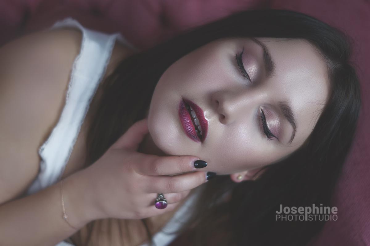 Portret kobiety z zamkniętymi oczami, delikatnym makijażem, burgundową szminką i pierścionkiem z fioletowym kamieniem, dłoń pod brodą, w tle różowa tkanina.