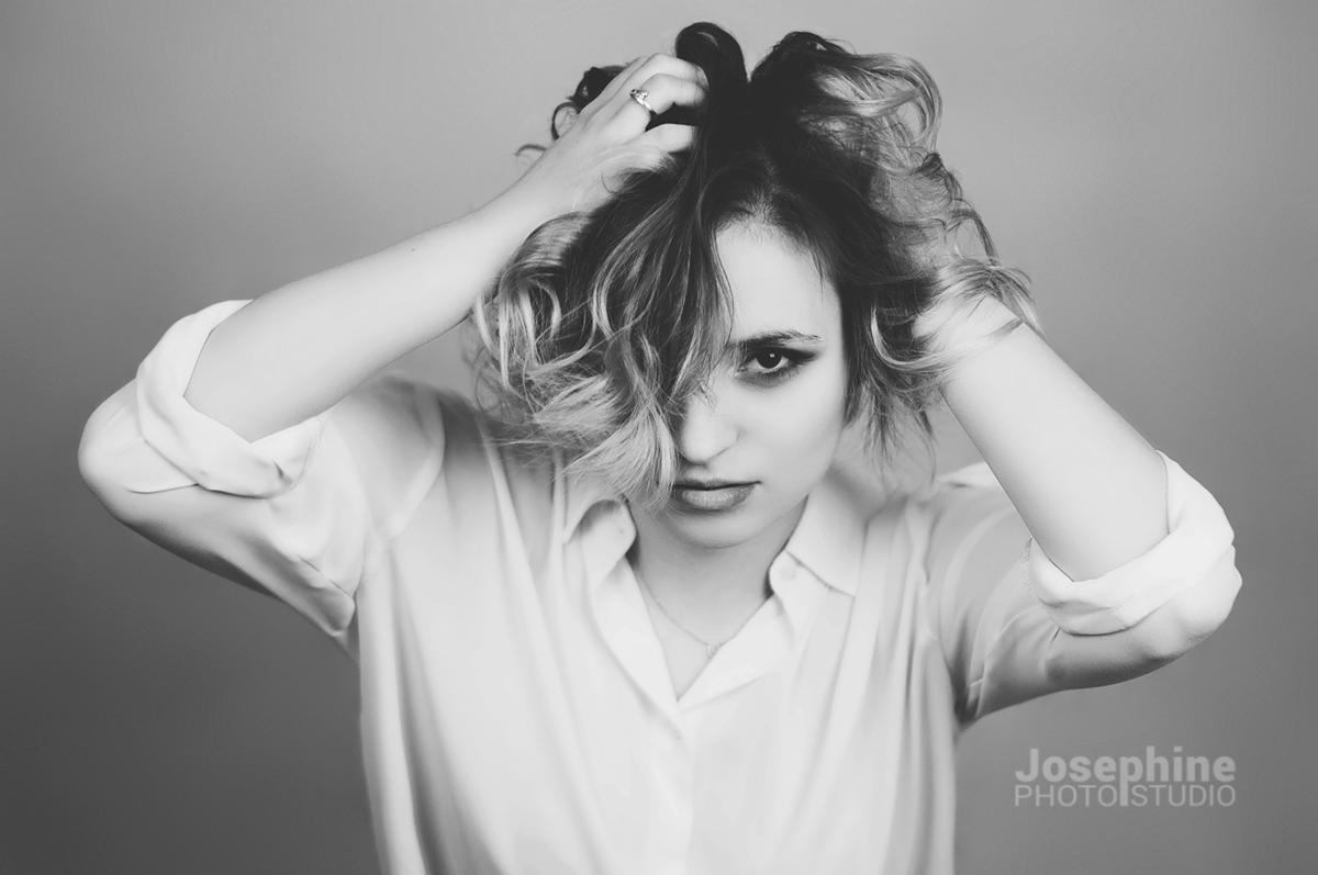 Czarno-białe portretowe ujęcie kobiety z kręconymi włosami, w białej koszuli, z rękami uniesionymi do góry i pierścionkiem na palcu, patrzącej wprost w obiektyw, z logo studia fotograficznego...