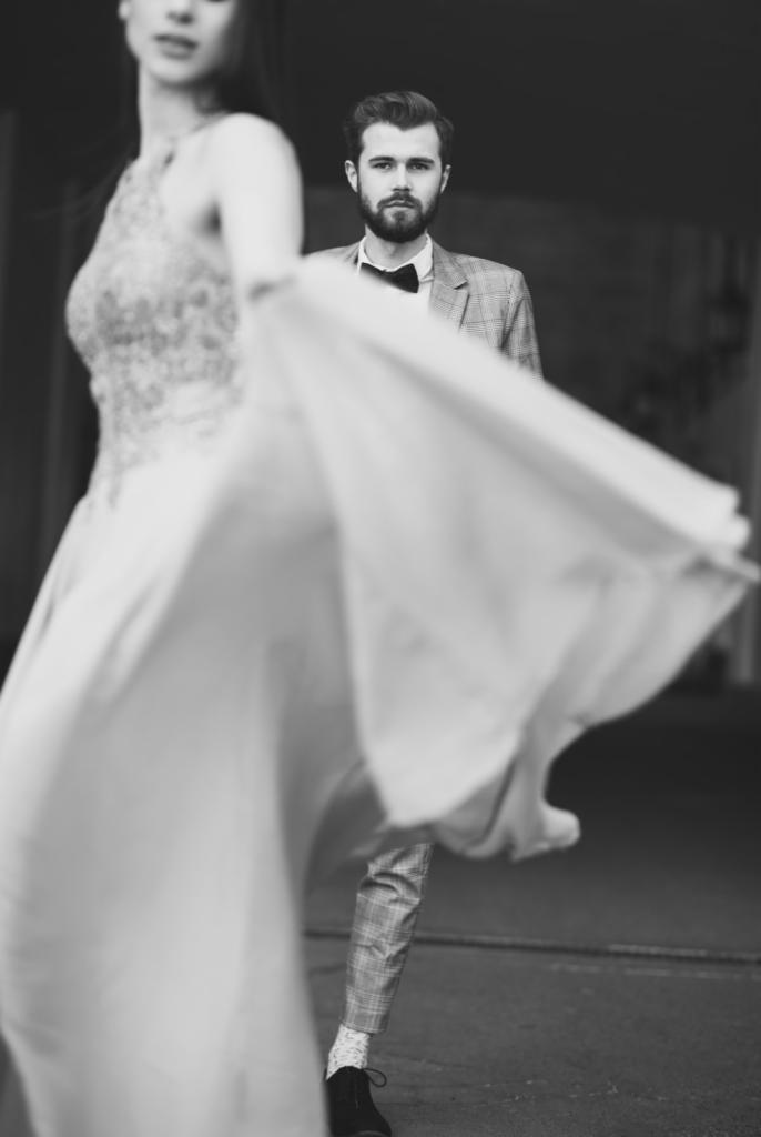 Czarno-białe zdjęcie pary: panna młoda w rozmytej sukni z falującym rękawem i pan młody w kraciastej marynarce i muszce, stojący w tle.