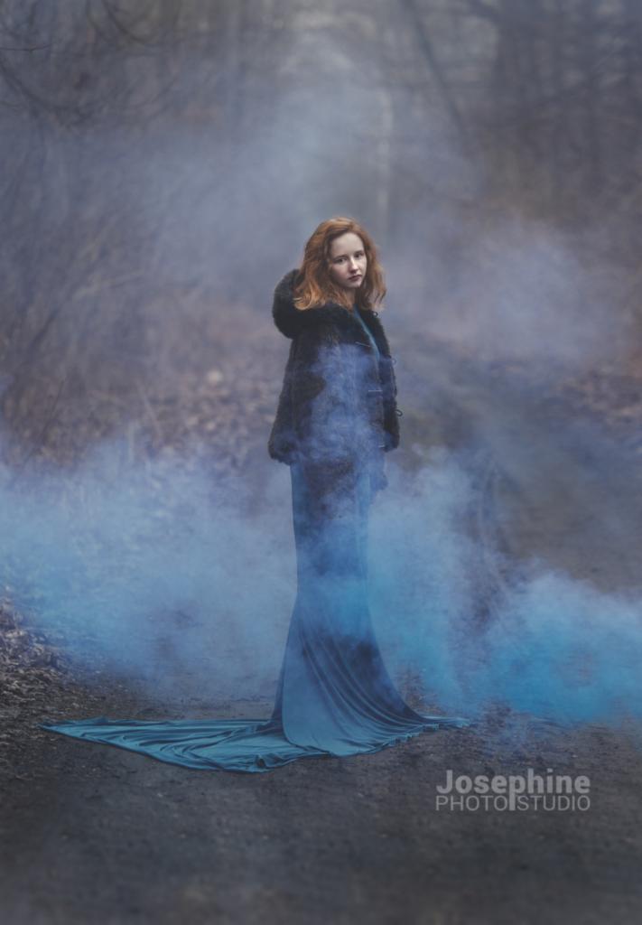 Rudowłosa kobieta w długiej turkusowej sukni i futrzanej kurtce z kapturem pozuje w lesie otoczona niebieską mgłą.
