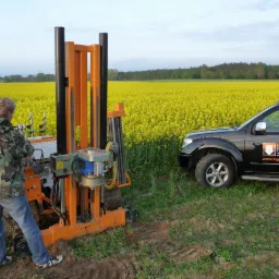Pracownik obsługuje wiertnicę geologiczną na gąsienicach na tle pola rzepaku, obok stoi czarny samochód terenowy z logo firmy.