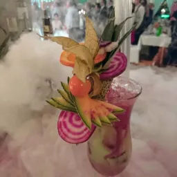 Egzotyczny koktajl z dekoracją z owoców i warzyw, otoczony suchym lodem na tle imprezy.