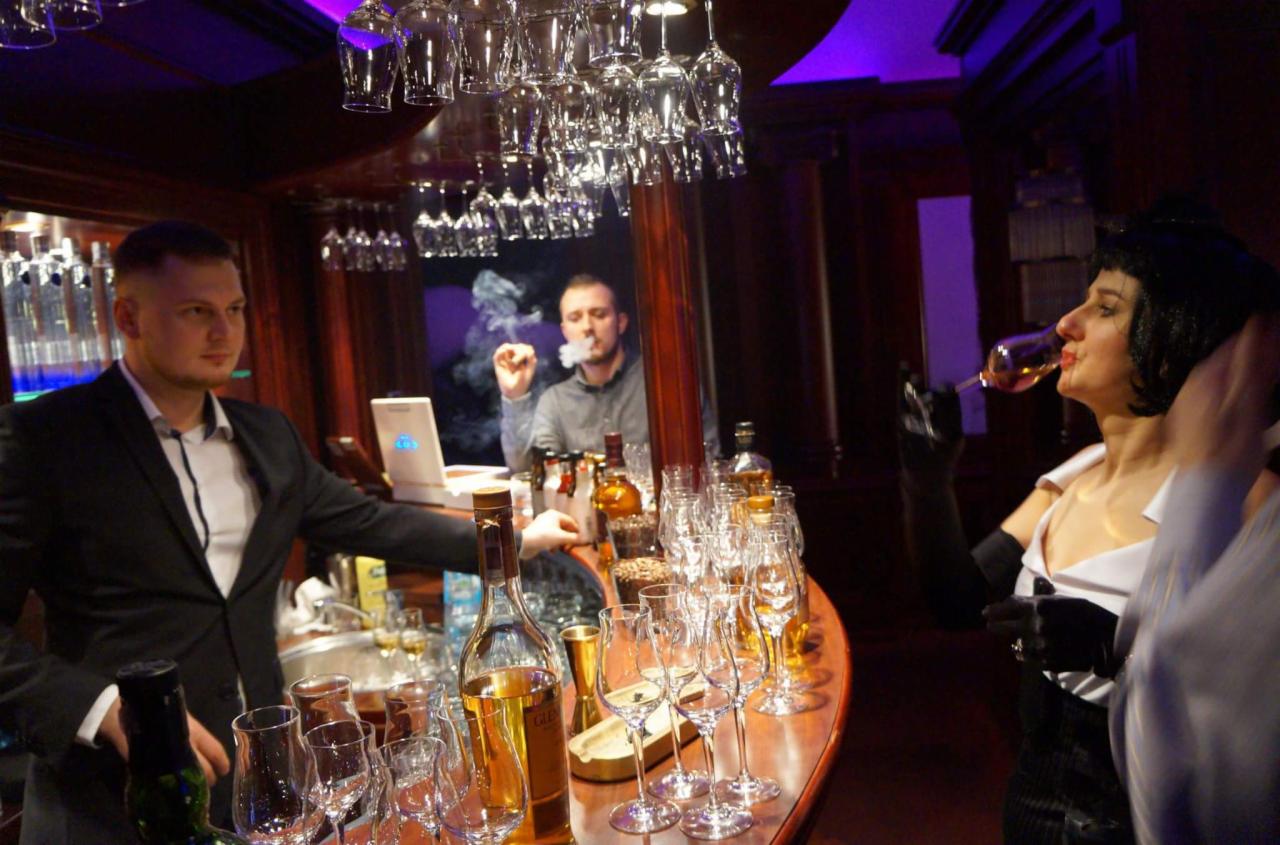 Elegancko ubrani ludzie przy barze z alkoholem, jeden mężczyzna w tle pali cygaro, na barze ustawione butelki i kieliszki.