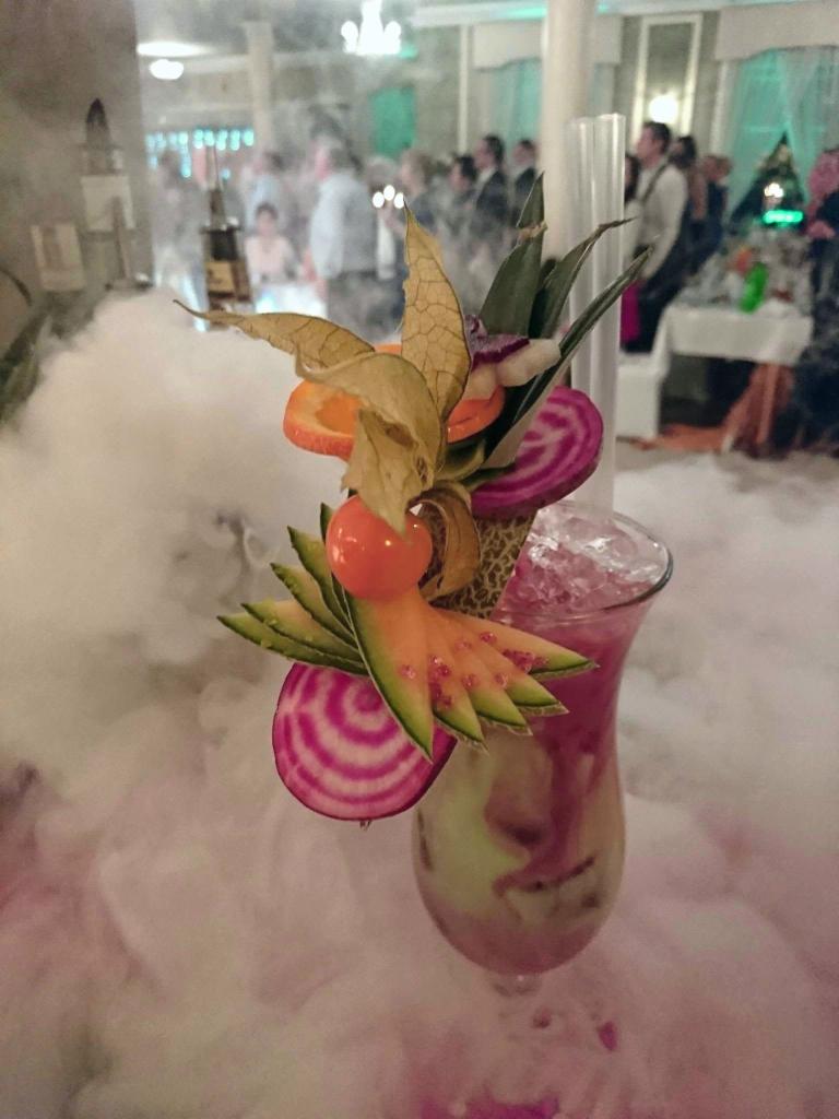 Egzotyczny koktajl z dekoracją z owoców i warzyw, otoczony suchym lodem na tle imprezy.