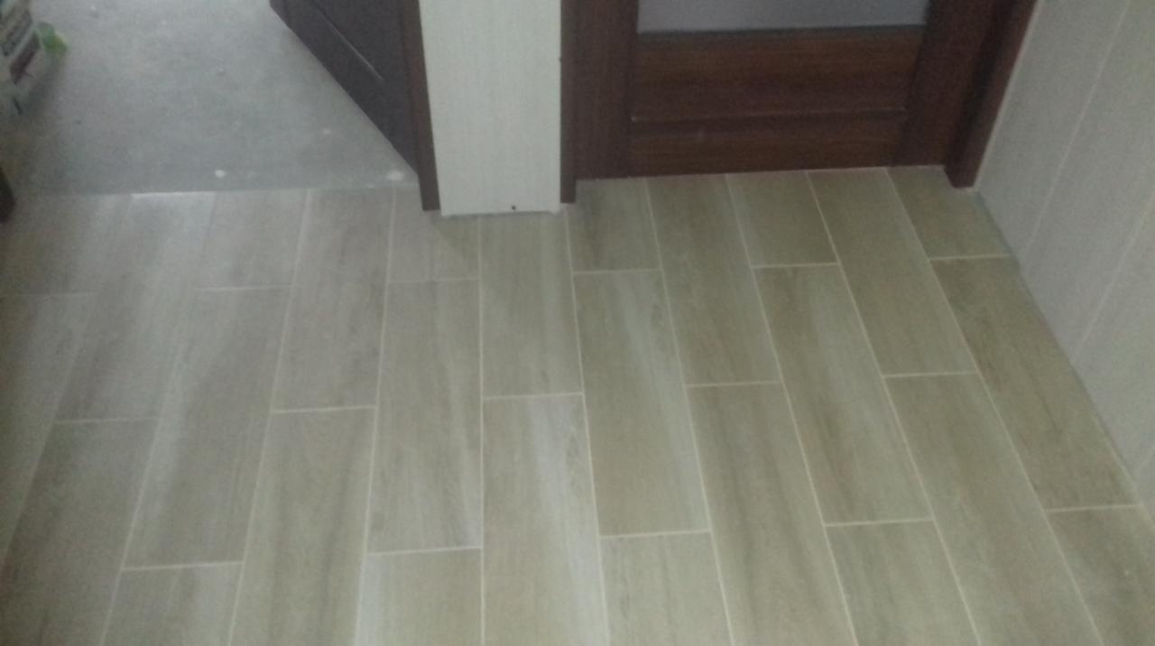 Ułożona podłoga z podłużnych, beżowych płytek imitujących drewno, widoczne fragmenty ściany i futryny drzwi.