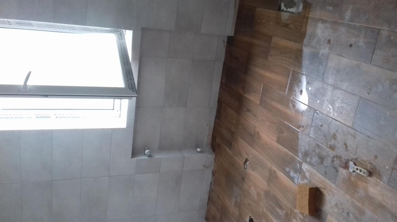 Montaż płytek imitujących drewno na podłodze łazienki, widoczne świeże ślady po kleju, gąbka i szpachelka, jasne płytki na ścianach i okno dachowe.