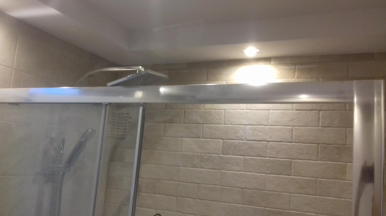 Kabina prysznicowa z deszczownicą i słuchawką, wykończona beżowymi płytkami imitującymi cegłę, oświetlona punktowymi lampami w suficie.