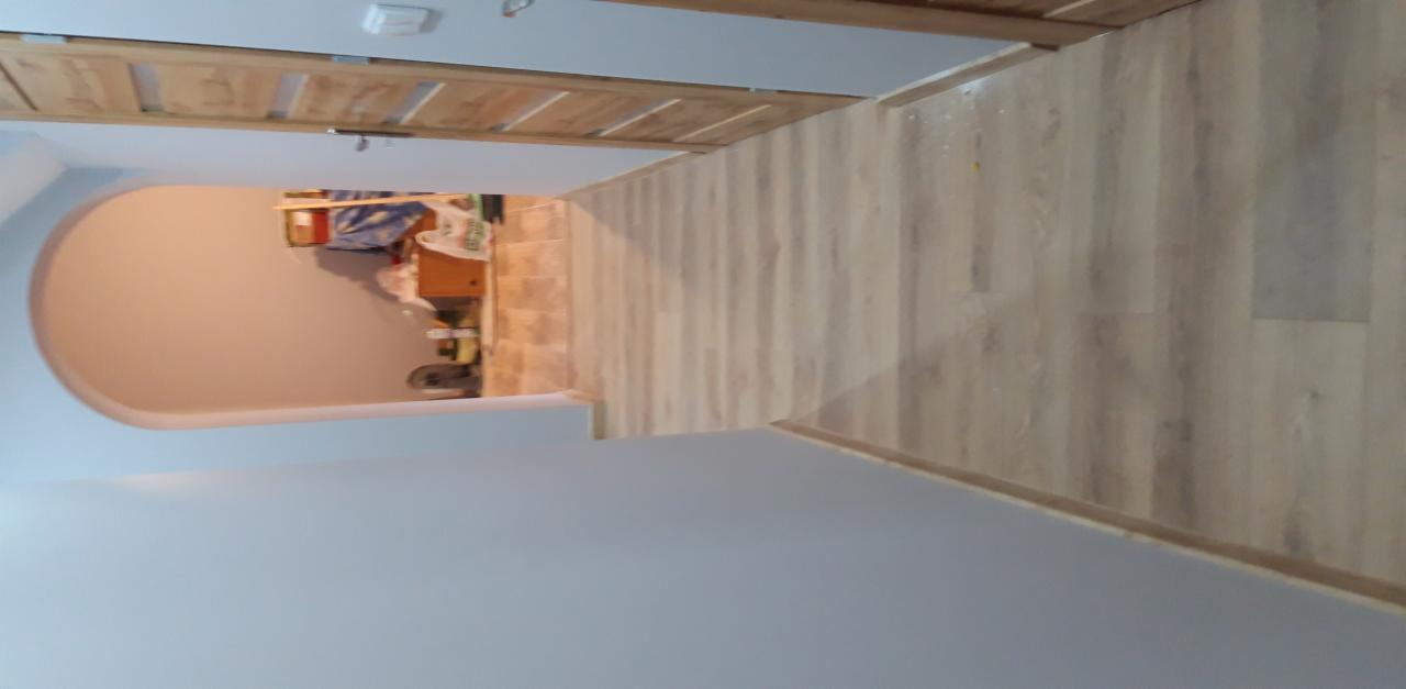 Ułożona podłoga z paneli drewnopodobnych w korytarzu, widok na łukowe przejście do innego pomieszczenia z widocznym wyposażeniem.