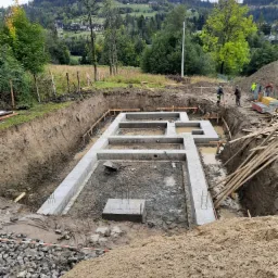 Wykop pod fundamenty domu w kształcie litery H, częściowo zalany wodą, z widocznymi zbrojeniami, drewnianymi szalunkami i pracującymi robotnikami na tle zalesionych wzgórz.