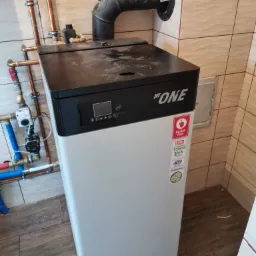 Nowoczesny piec grzewczy HT ONE z czarnym panelem sterowania i białym korpusem, zamontowany w pomieszczeniu z beżowymi płytkami, widoczne miedziane rury instalacji grzewczej.