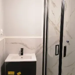 Nowoczesna łazienka z czarną szafką pod umywalką i kabiną prysznicową z czarnymi ramami, wykończona białymi płytkami imitującymi marmur, oświetlona okrągłą, szklaną lampą.