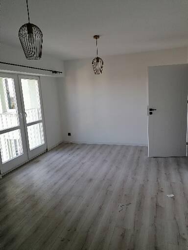 Puste, jasne pomieszczenie z szarymi panelami podłogowymi, białymi ścianami i drzwiami balkonowymi, dwie nowoczesne lampy wiszące z metalowymi kloszami.