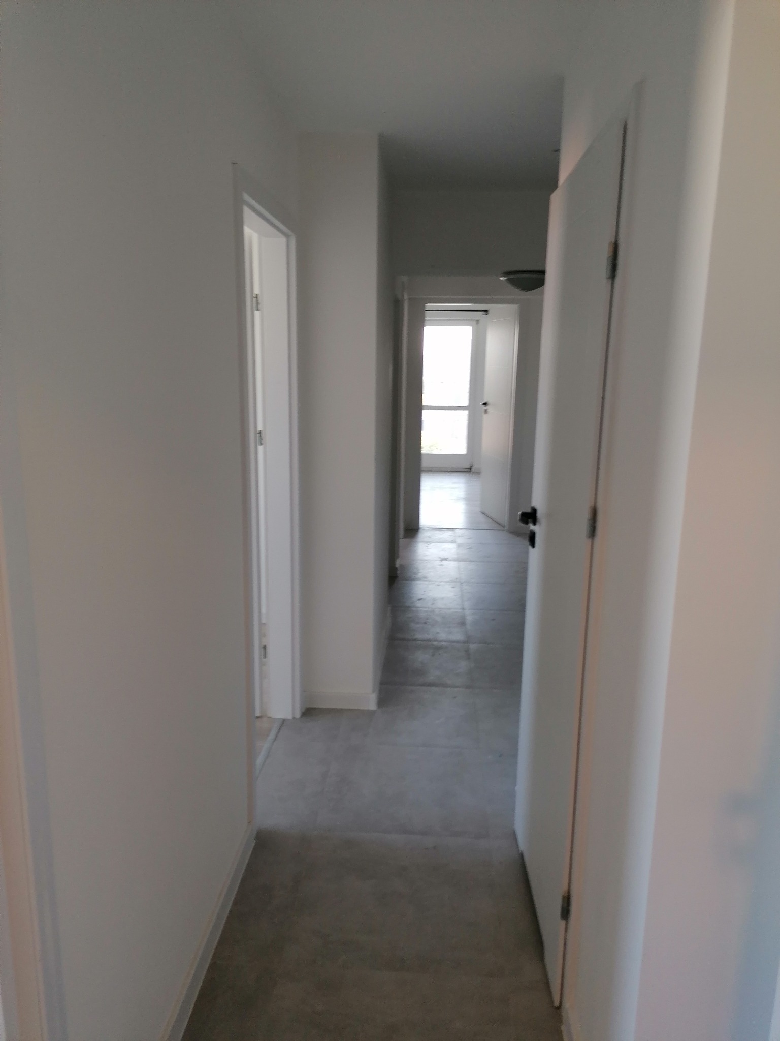 Wnętrze korytarza z szarymi płytkami podłogowymi i białymi ścianami, widok na otwarte drzwi na końcu korytarza, minimalizm w wykończeniu.