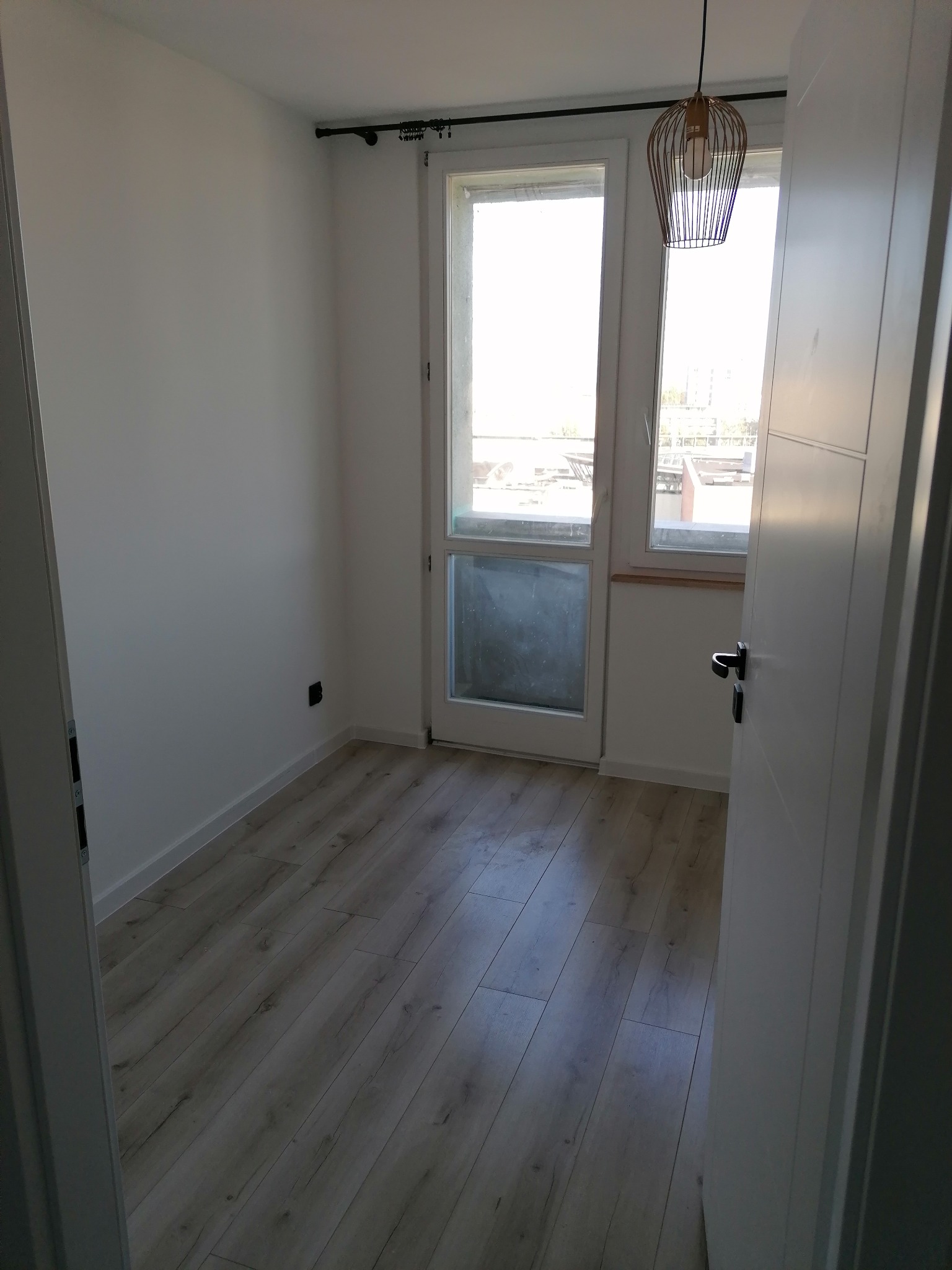 Pusty pokój z panelami podłogowymi w odcieniach szarości, białymi ścianami, oknem balkonowym i wiszącą lampą z ażurowym kloszem.